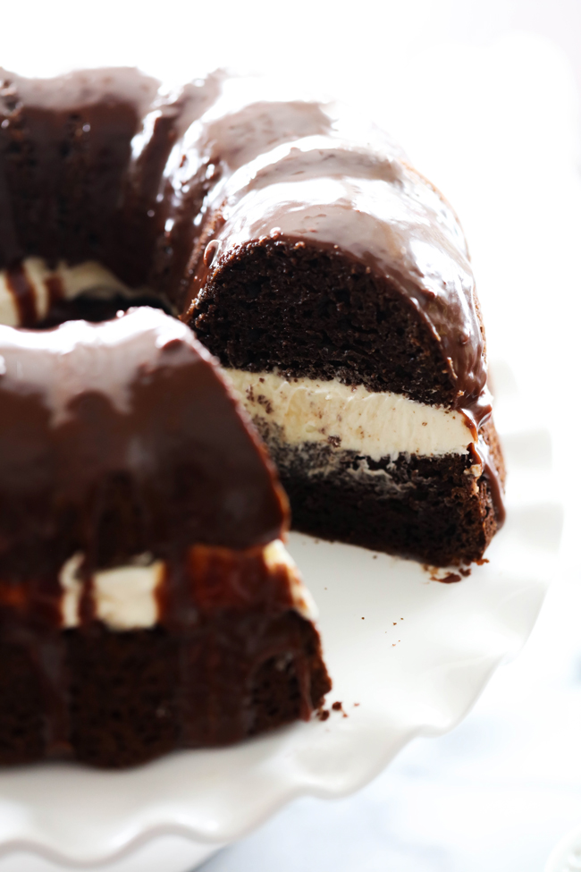 Ding Dong Bundt Cake is displayed on a white cake stand with slice removed to reveal layers of cake, filling and chocolate ganache.