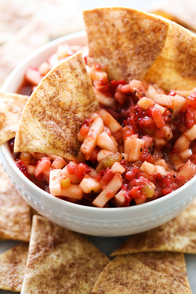 Three cinnamon sugar chips dipping into Fruit Salsa in a white bowl surrounded by cinnamon sugar chips.
