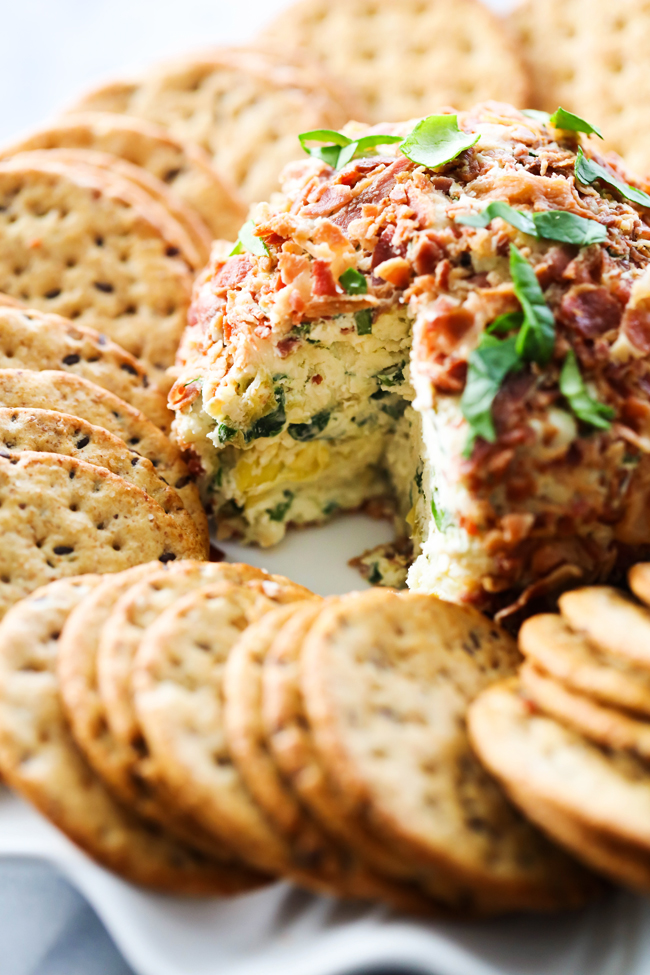 Spinach Artichoke Cheese Ball coated in bacon crumbs and garnished with fresh chopped spinach, with section removed to reveal middle, surrounded by crackers on white serving tray.