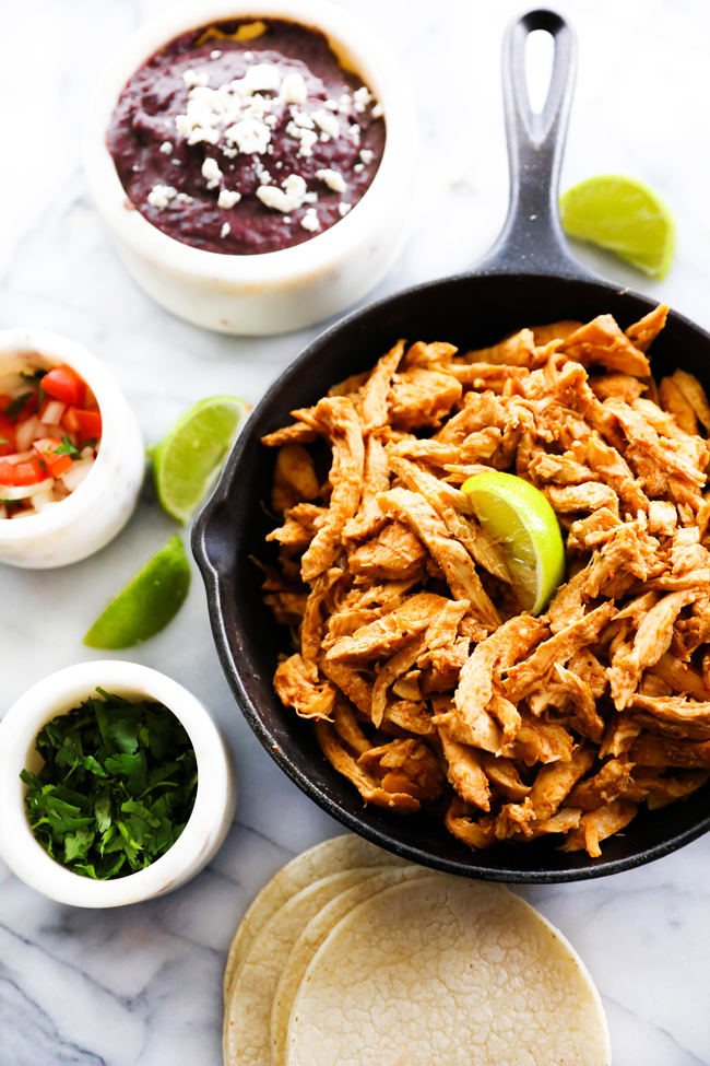 Chipotle Chicken in a small cast iron skillet surrounded by separate containers containing black beans, fresh chopped cilantro, pico de gallo, sliced limes and corn tortillas.