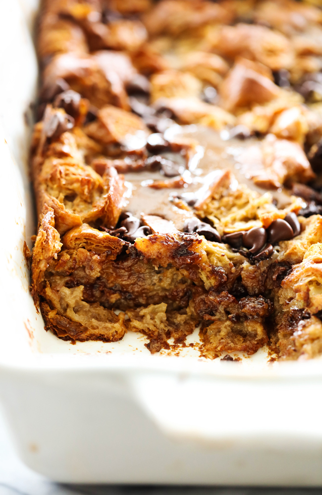 Chocolate Croissant Bread Pudding in a white casserole pan that is topped with syrup and a corner piece removed.