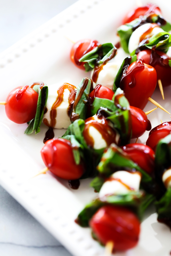 Cherry tomatoes, basil leaves and mozzarella balls skewered together and drizzled with balsamic glaze on-top a white plate.
