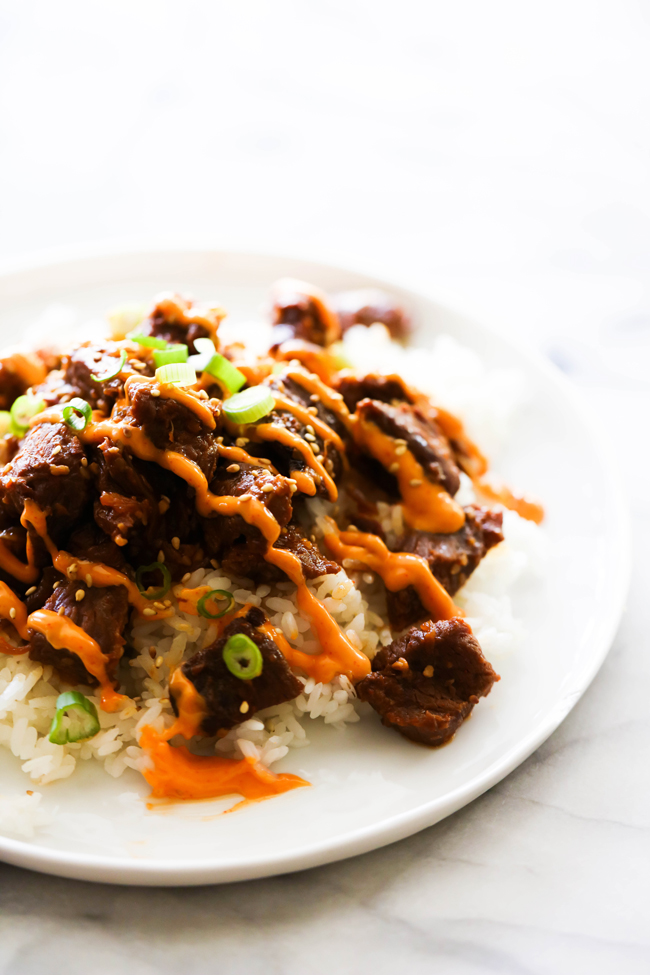 Korean Beef Bulgogi on a white plate with Gochujang Mayo drizzled over the top and garnished with green onions and sesame seeds.